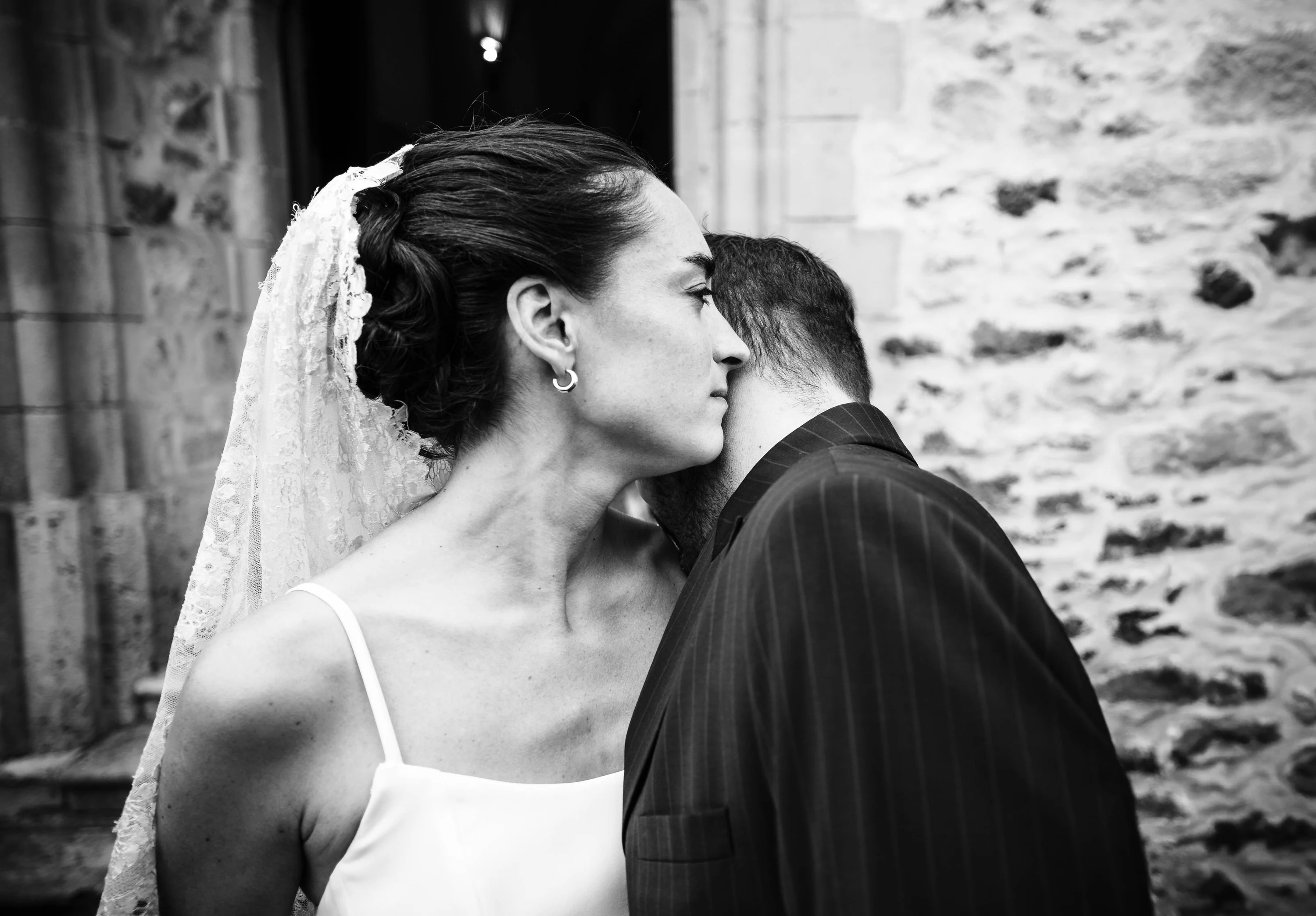 Noir et blanc : le marié embrasse la nuque de la mariée devant un mur de pierre